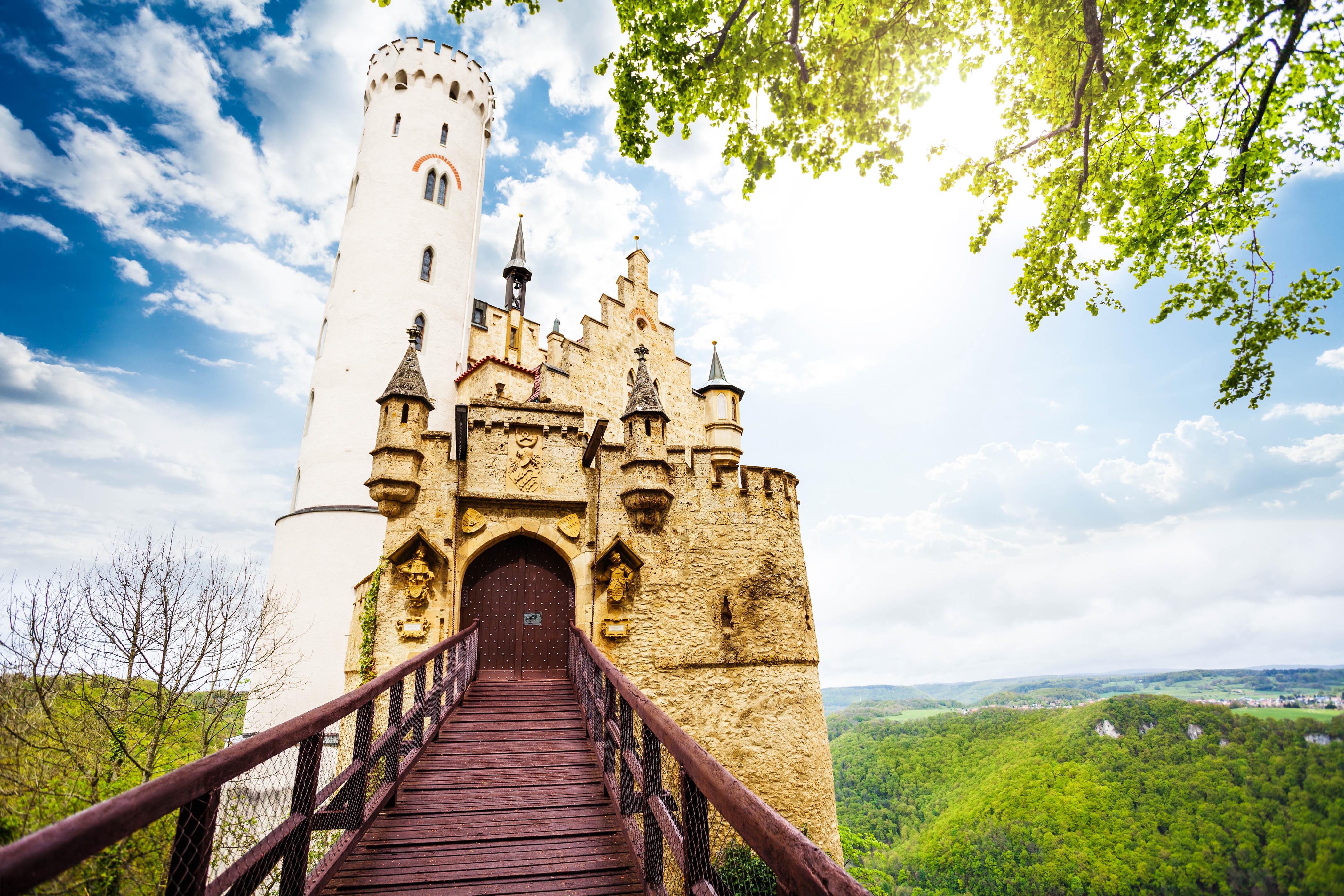 Holzbrücke führt zu Schloss Lichtenstein mit einem hohen, weißen Turm links. Rechts Wälder und Hügel unter blauem Himmel. Baumkrone oben rechts ragt ins Bild.