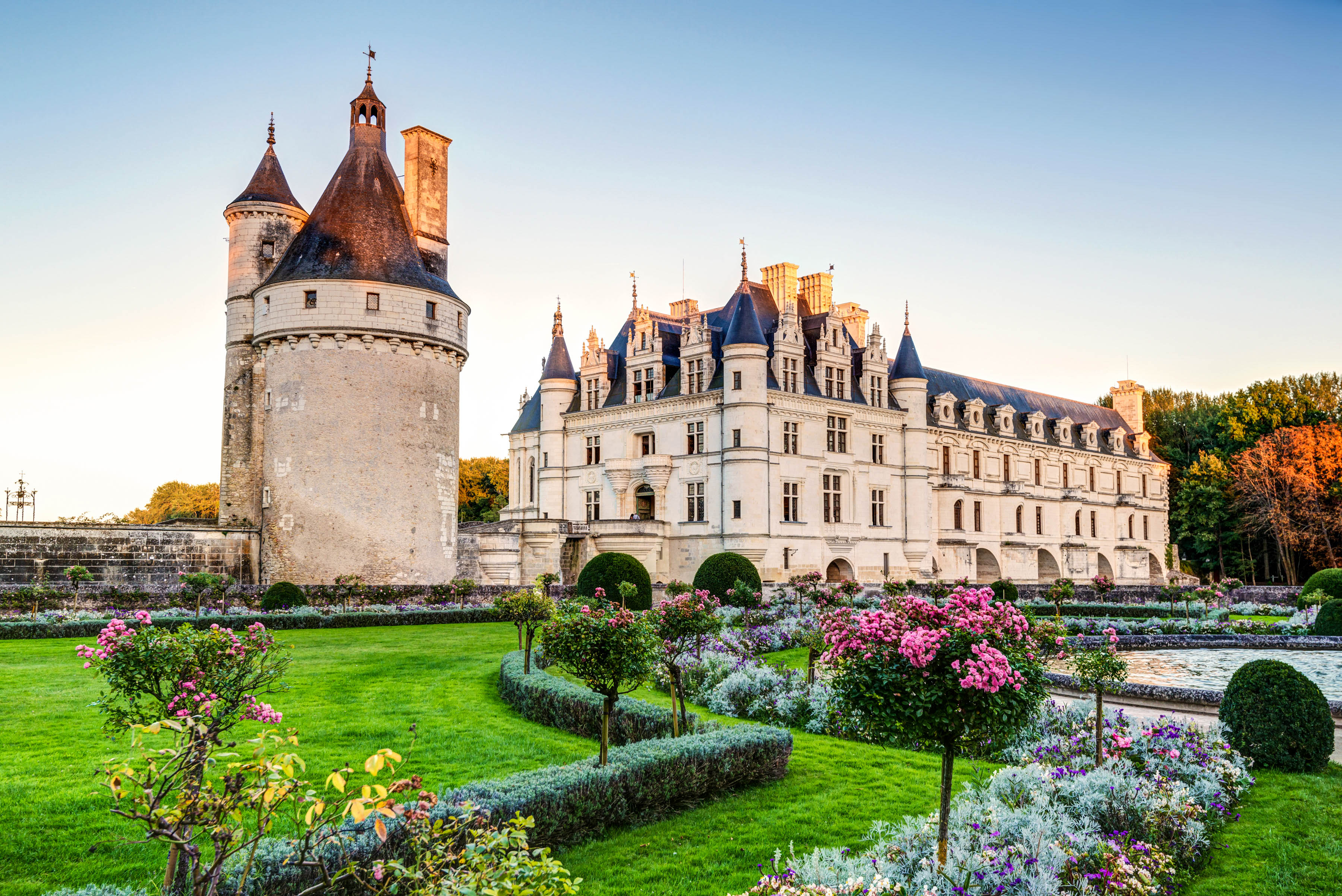 Schloss mit Türmen und Erkern, umgeben von gepflegten Gärten und bunten Blumenbeeten. Im Vordergrund grüner Rasen, im Hintergrund Bäume unter klarem Himmel bei Abenddämmerung.