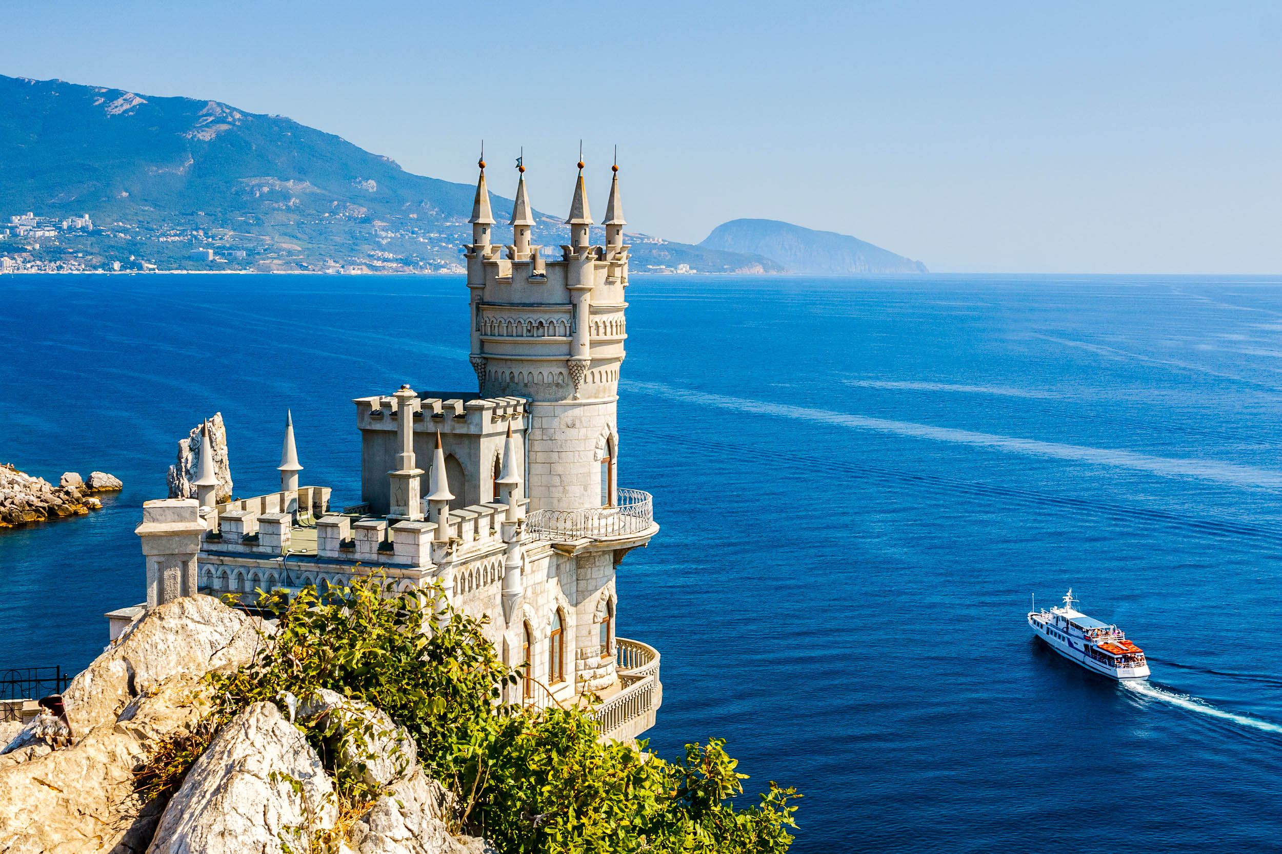 Märchenhaftes Schloss mit Türmen auf Klippe, Meer und Boot rechts, im Hintergrund grüne Berge. Klare, sonnige Atmosphäre und tiefblaues Wasser dominieren die Szenerie.