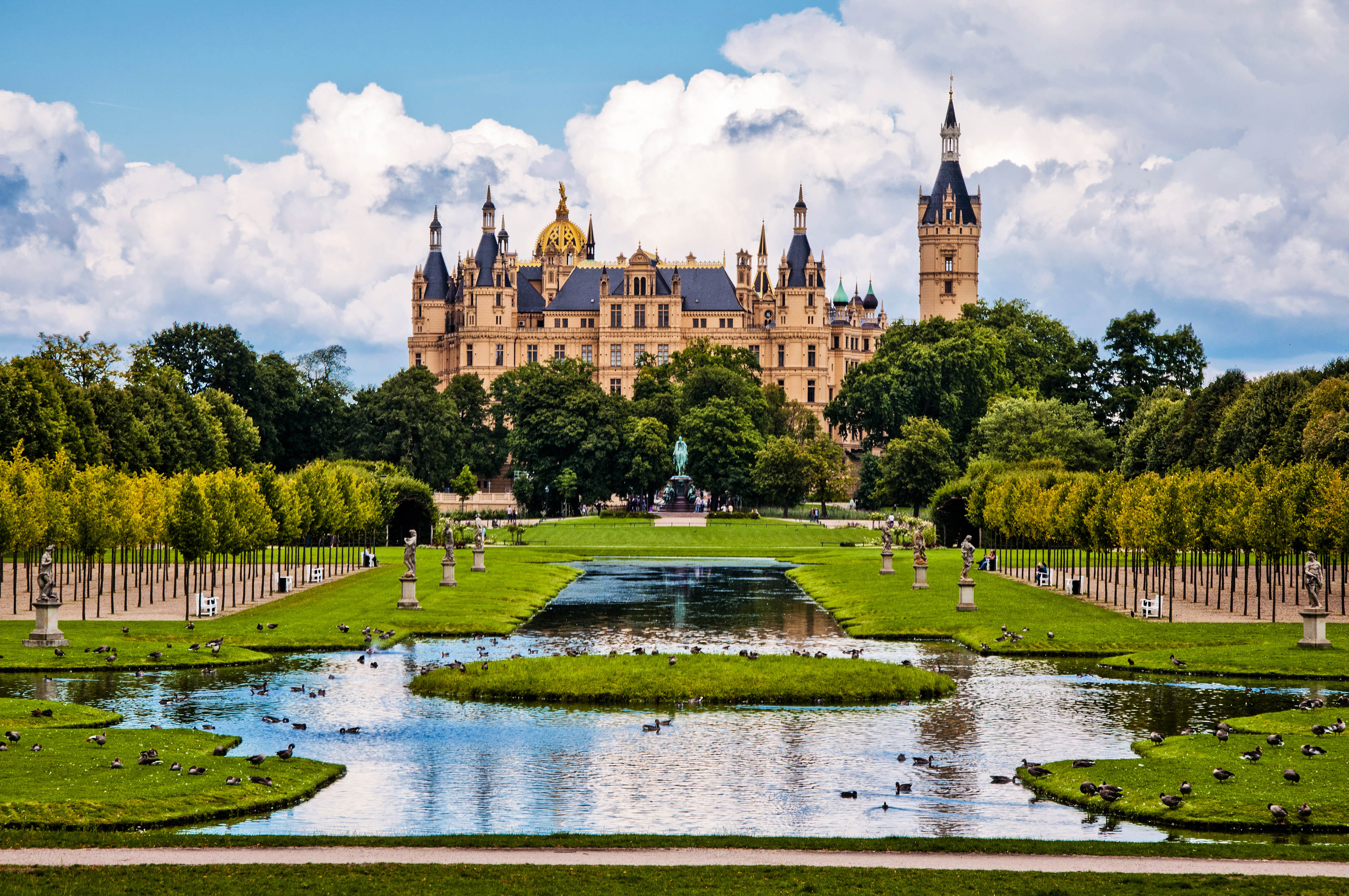 Märchenschloss mit goldener Kuppel und Türmen thront im Hintergrund über einer gepflegten Gartenanlage mit Bäumen und einem Teich voller Vögel; im Fokus eine Statue auf einem begrünten Weg.